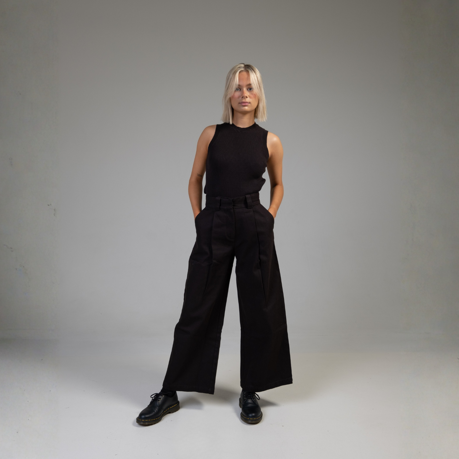 Female model wearing a black sleeveless top and wide-leg black trousers, posing in a minimalist studio for Nowa clothing brand product showcase.