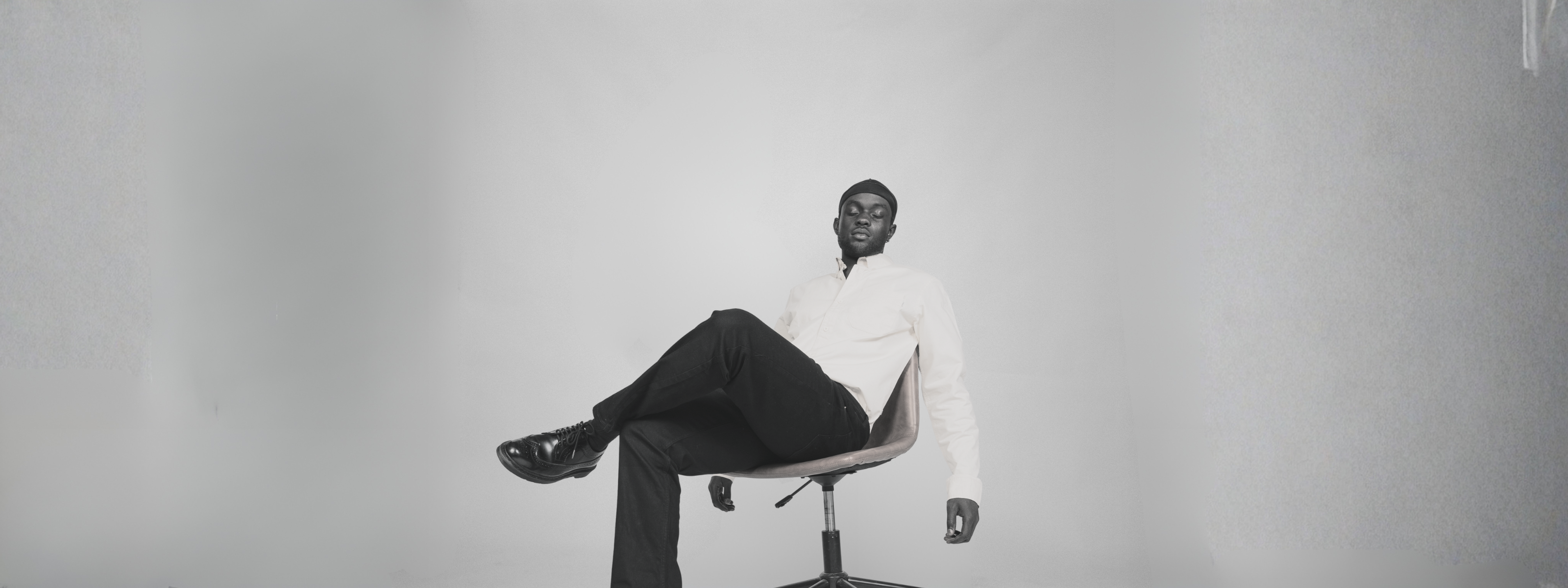 Male model wearing a minimalist light button-up shirt and black trousers, posing in a modern studio chair for Nowa clothing brand fashion shoot.