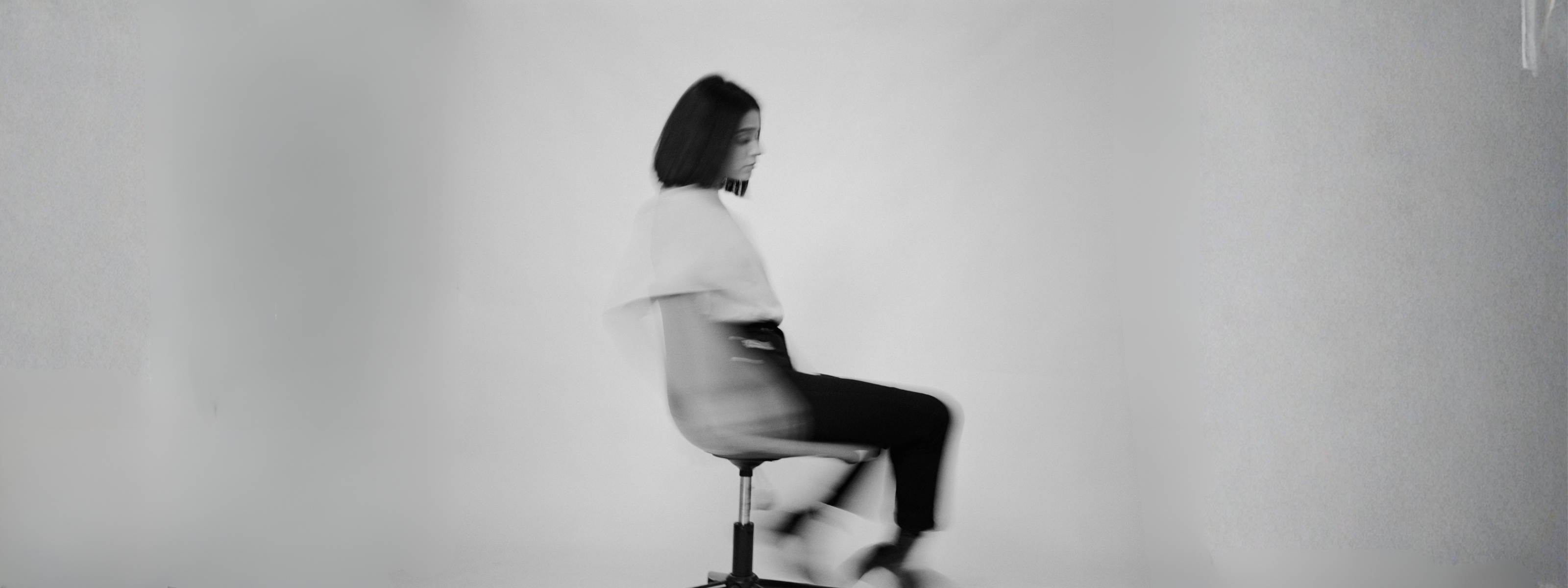 Female model in a minimalist white top and black trousers captured in motion on a studio chair for Nowa clothing brand campaign.