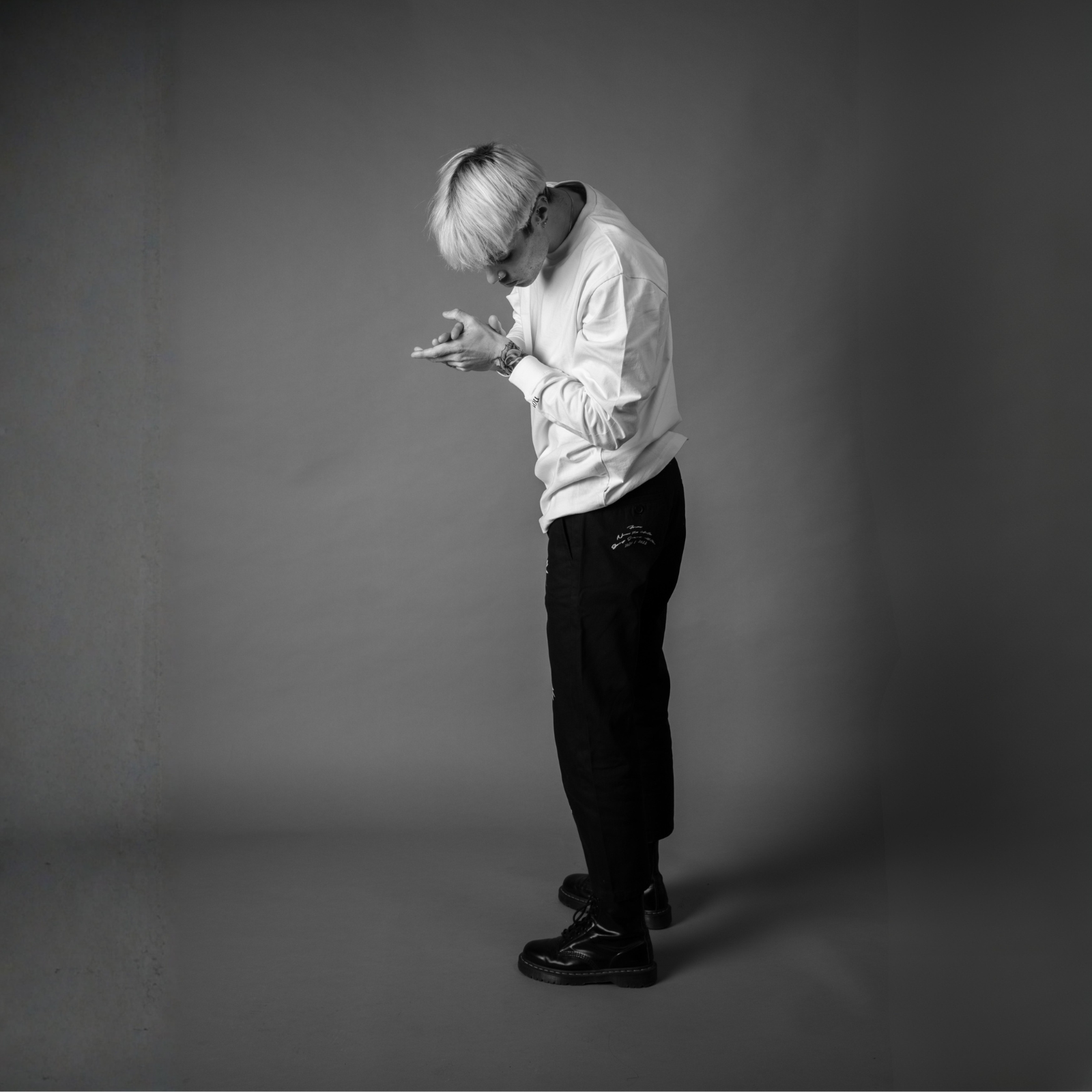 Minimalist black and white studio photo of a model with blonde hair wearing a white long-sleeve top, dark trousers, and polished shoes, posed looking down.